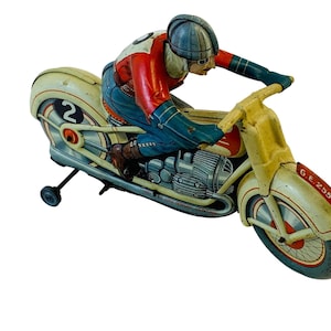 May include: A vintage tin toy motorcycle with a rider wearing a helmet and racing number 2. The motorcycle is painted white, yellow, red, and blue. The motorcycle has a G.E. 255 label on the side.