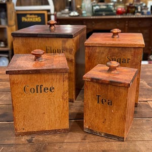 Puede incluir: Un juego de cuatro recipientes cuadrados de madera con tapas y pomos. Los recipientes tienen etiquetas con texto negro que dice Flour, Sugar, Coffee y Tea. Cada recipiente presenta un acabado de veta de madera natural con bordes oscuros.