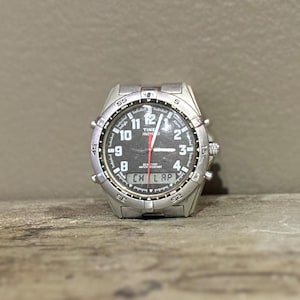 Peut inclure: Une montre Timex Indiglo argentée avec un cadran noir et un affichage numérique. La montre a un bracelet noir et une lunette argentée.