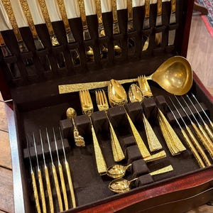 May include: A gold-toned flatware set housed in a dark wood storage case with a white satin-lined lid. The set includes knives, forks, spoons, and a large ladle, all featuring intricate embossed patterns on the handles and polished gold finishes.