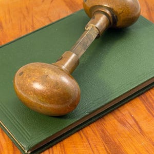 May include: An aged brass door knob with an oval design, placed on a green book. The door knob has a weathered look, indicating its vintage nature. The book is a deep green, and the background is a wooden surface.