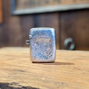 Puede incluir: Un porta-cerillas de plata con un diseño grabado ornamentado. El porta-cerillas tiene una tapa con bisagras y un pequeño lazo para llevarlo.