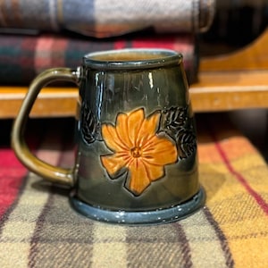 Puede incluir: Una taza de cerámica verde oscuro con un diseño de flor naranja en relieve. La taza tiene un asa y una base ancha.