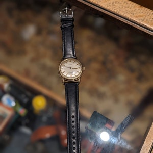 May include: A vintage Cyma wristwatch with a gold-toned case and a black leather strap. The watch face is light-coloured with gold hands and hour markers. The black leather strap has a buckle closure. The watch is displayed on a reflective surface.