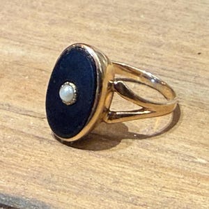 Peut inclure: Bague dor&eacute;e orn&eacute;e d'une pierre ovale bleu fonc&eacute; avec une petite perle blanche ronde au centre. La bague a un design &agrave; tige fendue. La bague est pos&eacute;e sur une surface en bois.