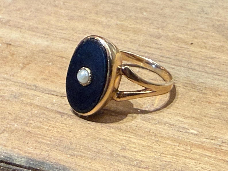 Peut inclure: Bague dor&eacute;e orn&eacute;e d'une pierre ovale bleu fonc&eacute; avec une petite perle blanche ronde au centre. La bague a un design &agrave; tige fendue. La bague est pos&eacute;e sur une surface en bois.