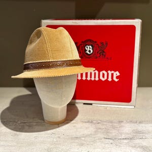 Op de afbeelding: Een bruine corduroy fedora hoed met een bruine leren band wordt getoond op een mannequin hoofd. De hoed staat voor een rode doos met het woord "Baltimore" in het wit.