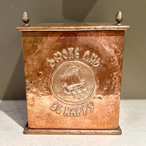 Può includere: Una scatola di rame con finitura martellata e dettagli in ottone. La scatola ha un'iscrizione in rilievo che recita "Smoke and Be Happy" e presenta un design di una nave.