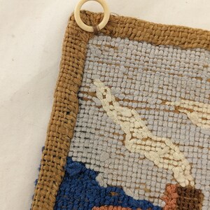 Op de afbeelding: Een close-up van een geweven wandkleed met een bruine jute rand. Het wandkleed heeft een kleurrijk ontwerp met tinten blauw, groen, geel en oranje. Een witte plastic ring is bevestigd aan de bovenkant van het wandkleed.
