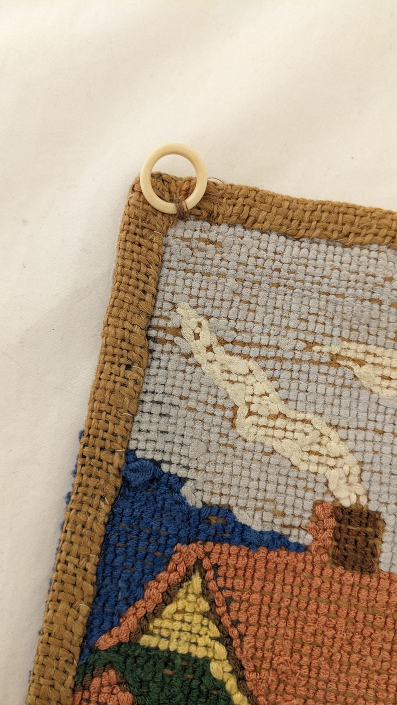 Op de afbeelding: Een close-up van een geweven wandkleed met een bruine jute rand. Het wandkleed heeft een kleurrijk ontwerp met tinten blauw, groen, geel en oranje. Een witte plastic ring is bevestigd aan de bovenkant van het wandkleed.