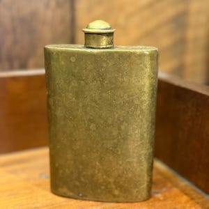 May include: An antique brass flask, rectangular in shape with a rounded top and a small, screw-on cap. The flask has a slightly textured surface and a tarnished, aged appearance. The cap is a lighter shade of brass with a ridged edge.