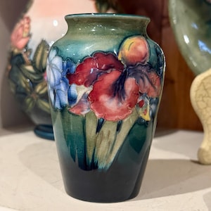 Puede incluir: Un pequeño jarrón de cerámica con una base verde oscuro que se desvanece a un verde más claro en la parte superior. El jarrón está decorado con diseños florales rojos, azules y amarillos. El jarrón probablemente se utiliza con fines decorativos.