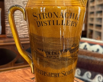 Puede incluir: Una jarra de cerámica esmaltada en un tono amarillo mostaza cálido con base y borde de color marrón oscuro. El frente presenta texto negro que dice STRONACHIE DISTILLERY y PERTHSHIRE SCOTLAND, junto con una detallada ilustración negra de una destilería.