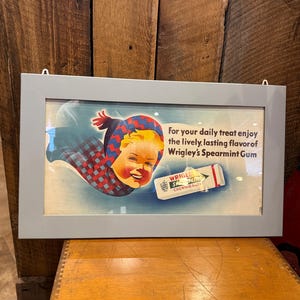 May include: Framed vintage advertisement for Wrigley's Spearmint Gum. The artwork features a smiling child wearing a blue and red plaid hat and scarf. Text reads, "For your daily treat enjoy the lively, lasting flavor of Wrigley's Spearmint Gum."