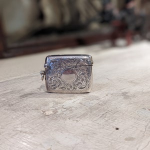May include: A silver match safe with an ornate engraved design. The match safe has a hinged lid and a small loop for attaching a chain.