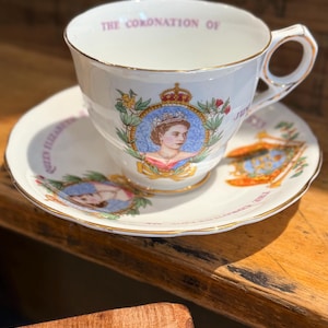 Op de afbeelding: Een witte theekop en schotel set met gouden rand. De kop en schotel tonen een portret van koningin Elizabeth II, een kroon en de tekst "THE CORONATION OF". De set is waarschijnlijk een herdenkingsartikel.