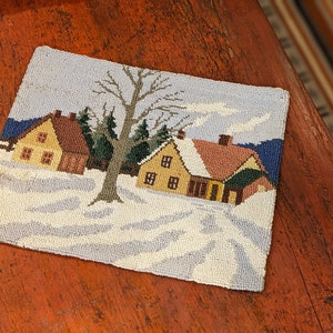 Op de afbeelding: Een naaldkunst wanddecoratie met een wintersc&egrave;ne met twee huizen en een kale boom op de voorgrond. De huizen zijn geel en bruin met witte sneeuw op de daken. De achtergrond is een blauwe lucht met witte wolken.