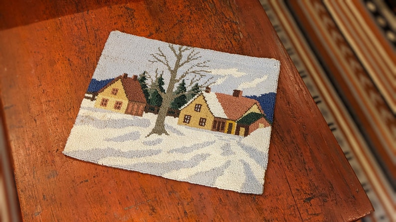 Op de afbeelding: Een naaldkunst wanddecoratie met een wintersc&egrave;ne met twee huizen en een kale boom op de voorgrond. De huizen zijn geel en bruin met witte sneeuw op de daken. De achtergrond is een blauwe lucht met witte wolken.