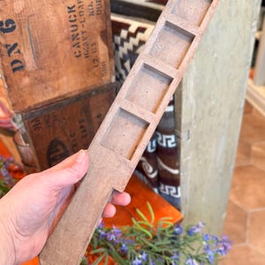 May include: A light brown, wooden tool with four square indentations and a handle. The tool is held in a person's hand. In the background are wooden crates with text and purple flowers.