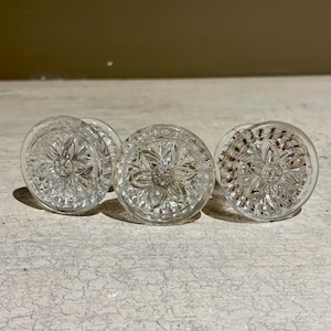 Può includere: Tre pomelli in vetro trasparente con un motivo floreale decorativo. I pomelli hanno un design circolare rialzato con un punto centrale.