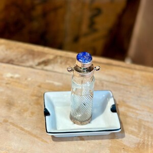 Könnte beinhalten: Eine kleine Glasflasche mit einem silbernen Verschluss und einem blauen Edelstein-Deckel. Die Flasche steht auf einer weißen Keramik-Schale mit schwarzem Rand.