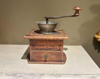 May include: Antique wooden coffee grinder with a metal hopper and a wooden crank handle. The grinder has a drawer for collecting coffee grounds and a label that reads "The New York Coffee Mill Co."