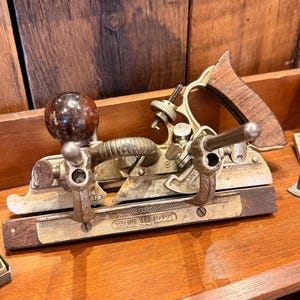 Puede incluir: Un cepillo de mano de carpintería vintage con mango de madera y cuerpo metálico. El cepillo tiene una cuchilla curva y está diseñado para dar forma a la madera. La herramienta es de metal y madera y se exhibe sobre una superficie de madera.