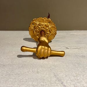 Könnte beinhalten: Goldfarbener Türklopfer mit einer Faust, die eine horizontale Stange hält. Der Türklopfer hat eine dekorative kreisförmige Rückplatte mit detailliertem Reliefdesign. Die Hardware ist aus Metall.