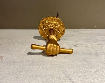May include: A gold-colored door knocker featuring a fist holding a horizontal bar. The knocker has a decorative circular backplate with a detailed relief design. The hardware is made of metal and is likely for a front door.