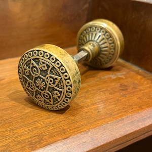 Puede incluir: Pomo de puerta antiguo de latón con un intrincado diseño circular. El pomo presenta un patrón detallado de formas geométricas y elementos florales. El latón tiene una pátina desgastada, lo que aumenta su atractivo vintage. El pomo está montado sobre una superficie de madera.