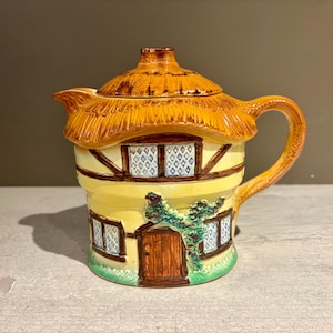 May include: A ceramic teapot shaped like a cottage. The roof is a thatched design, and the body is painted yellow with brown trim and windows. A green vine with leaves decorates the front. The handle and spout are curved.