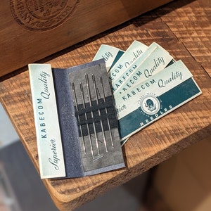 May include: A pack of needles labeled "Kabecorn Superior Quality" with a blue and green color scheme. The needles are in a black plastic case with a silver metal needle holder. The pack is sitting on a wooden surface.
