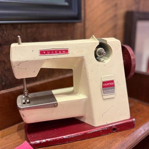 May include: A vintage off-white and red Vulcan Countess sewing machine. The machine has a red base and a cream-coloured body with the name "Vulcan" in red lettering. Made in England.