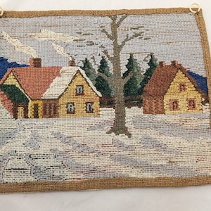 Op de afbeelding: Een vintage naaldkunst wandtapijt met een wintersc&egrave;ne met twee huizen, een boom en sneeuw. De huizen zijn geel en bruin met rode daken. De boom is groen en bruin. De sneeuw is wit en blauw.