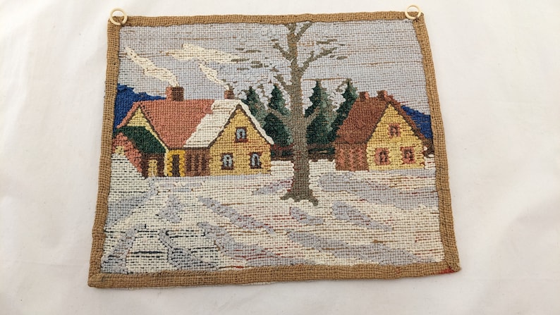 Op de afbeelding: Een vintage naaldkunst wandtapijt met een wintersc&egrave;ne met twee huizen, een boom en sneeuw. De huizen zijn geel en bruin met rode daken. De boom is groen en bruin. De sneeuw is wit en blauw.