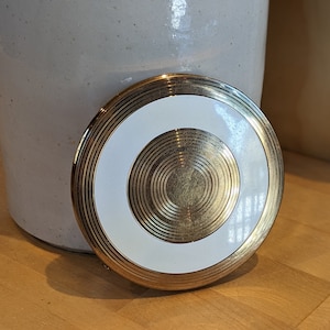 Op de afbeelding: Een goudkleurige compactspiegel met een witte cirkel in het midden. De cirkel heeft concentrische gouden ringen eromheen.