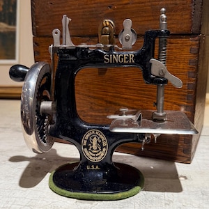 Puede incluir: Una máquina de coser Singer vintage de hierro fundido negro con manivela plateada, placa de aguja de metal y un emblema circular que dice The Singer Manfg. Co. U.S.A. La máquina presenta el logo de Singer en el brazo y una base de fieltro verde.