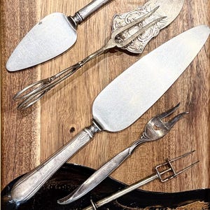 May include: A collection of five silver-toned serving utensils including two cake servers with smooth blades, a decorative serving fork with an ornate handle, a small two-pronged fork, and a slender metal pick with a sliding rectangular frame.