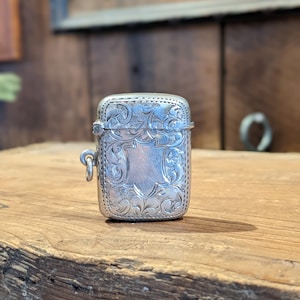 Puede incluir: Un porta-cerillas de plata con un diseño floral ornamentado. El porta-cerillas tiene una tapa con bisagras y un pequeño lazo para sujetar una cadena.