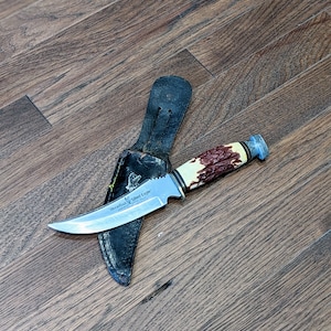 May include: A silver knife with a brown and white handle and a black leather sheath. The knife has the words "Woodland Silver Eagle" engraved on the blade.
