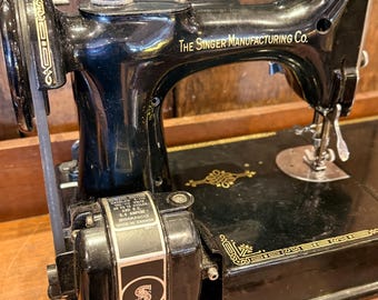 May include: A vintage black Singer sewing machine with gold accents. The machine has the text "The Singer Manufacturing Co." and a motor with the Singer logo. The machine is on a wooden surface.