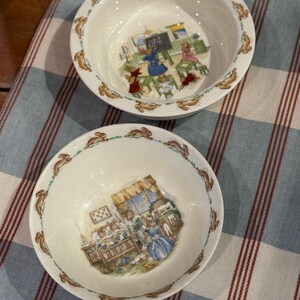 May include: Two white ceramic bowls with a rim of brown rabbits. The bowls feature illustrations of rabbits in a classroom and a bedroom. The bowls are on a blue and white striped tablecloth.