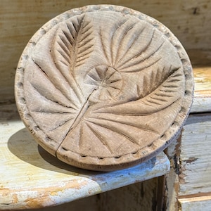 Könnte beinhalten: Eine runde Butterform aus hellbraunem Holz mit einem geschnitzten botanischen Motiv, das eine zentrale Blume mit strahlenden Blütenblättern und zwei flankierenden Blättern zeigt. Der kreisförmige Rand ist mit kleinen, gleichmäßigen Einkerbungen verziert.
