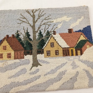 Op de afbeelding: Een naaldviltkussen met een wintersc&egrave;ne met drie huizen, een grote boom en een met sneeuw bedekte grond. De huizen zijn geel, bruin en rood met witte versieringen. De boom is bruin met groene bladeren. De sneeuw is wit en blauw.