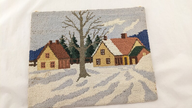 Op de afbeelding: Een naaldviltkussen met een wintersc&egrave;ne met drie huizen, een grote boom en een met sneeuw bedekte grond. De huizen zijn geel, bruin en rood met witte versieringen. De boom is bruin met groene bladeren. De sneeuw is wit en blauw.