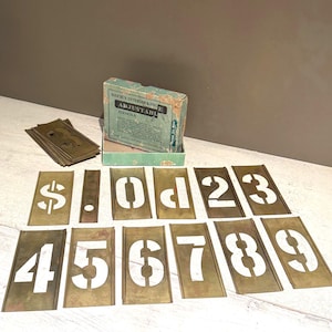 Peut inclure: Ensemble de pochoirs vintage avec des chiffres et symboles blancs sur des plaques métalliques couleur laiton. La boîte est étiquetée "Reese's Interlocking Adjustable Stencils".