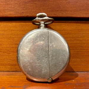 Könnte beinhalten: Eine silberne Taschenuhr mit einem runden Zifferblatt und einem Scharnier auf der Rückseite. Die Uhr hat ein schlichtes Design ohne sichtbare Markierungen oder Verzierungen.