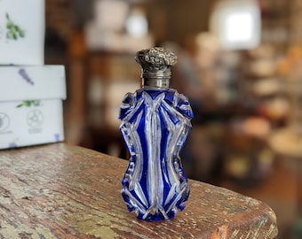 May include: A small, cobalt blue glass bottle with a silver stopper. The bottle has a faceted design and is sitting on a rustic wooden surface.