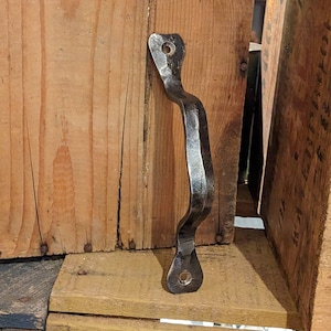 Op de afbeelding: Een handgesmede, donkere metalen handgreep met een gebogen ontwerp, gemonteerd op een houten oppervlak. De handgreep heeft een getextureerd uiterlijk en heeft gaten aan elk uiteinde voor montage. De achtergrond omvat houten kisten.
