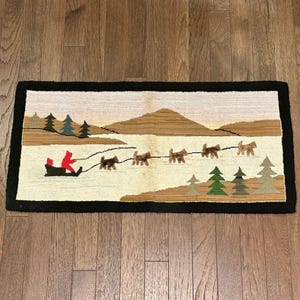 Könnte beinhalten: Rechteckiger Teppich mit einer Winterszene. Eine Person in einem roten Mantel fährt mit einem Schlitten, der von Hunden über eine verschneite Landschaft gezogen wird. Der Hintergrund zeigt Berge, Bäume und einen hellen Himmel. Der Teppich hat einen schwarzen Rand.